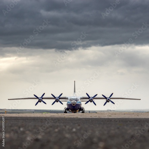 Front view of four propeller airplane. Big heavy gross weight airliner standing on airport apron, dynamic dark grey cloudy sky background. Huge freight aircraft for air cargo transportation.