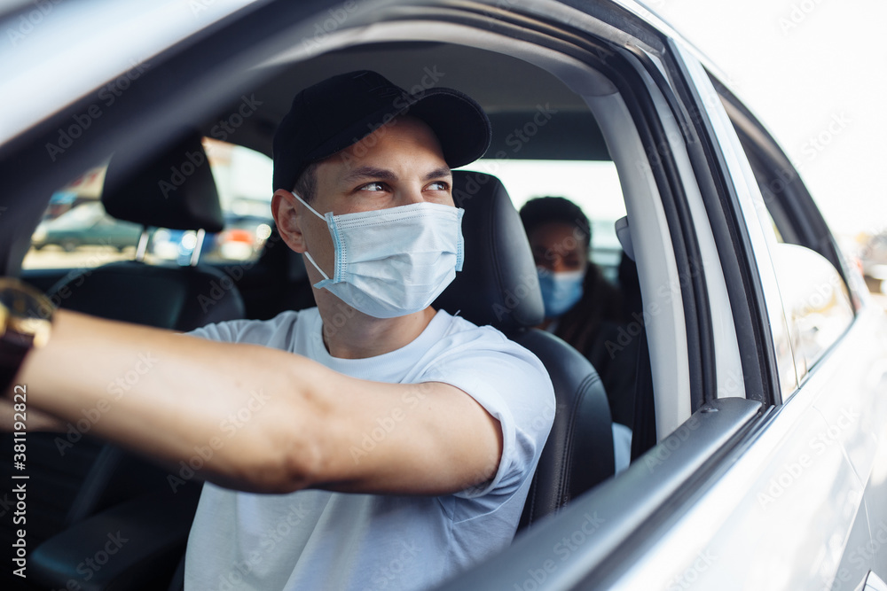 Young taxi driver looks out of a car's window while driving through the city with a passanger wearing a medical mask. Business trips during pandemic, new normal and coronavirus travel safety concept.
