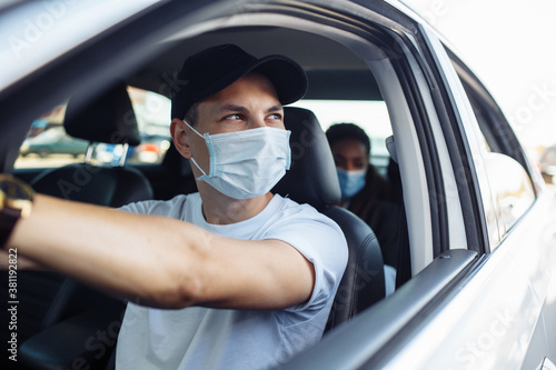 Young taxi driver looks out of a car's window while driving through the city with a passanger wearing a medical mask. Business trips during pandemic, new normal and coronavirus travel safety concept. © Konstantin Zibert