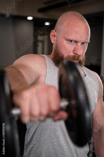 a strong athletic man with a red beard and a shaved head trines his arms with a dumbbell