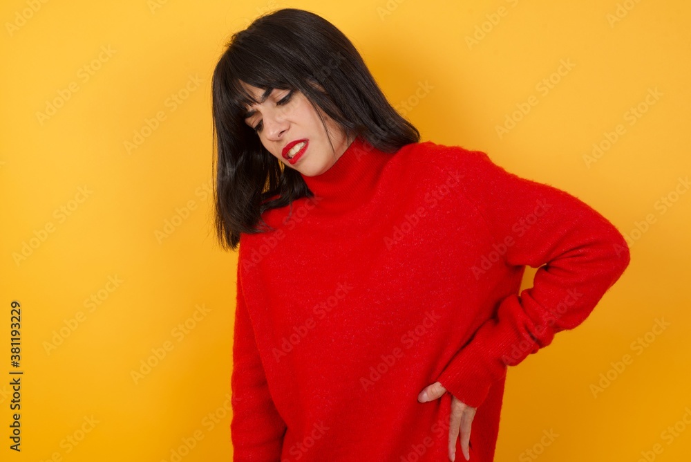 Caucasian brunette woman wearing red casual sweater isolated over yellow background Suffering of backache, touching back with hand, muscular pain Caucasian brunette woman wearing red casual sweater isolated over yellow background Suffering of backache, touching back with hand, muscular pain