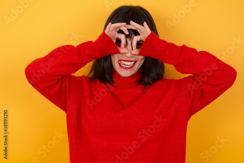 Caucasian brunette woman wearing red casual sweater isolated over yellow background doing ok gesture like binoculars sticking tongue out, eyes looking through fingers. Crazy expression.