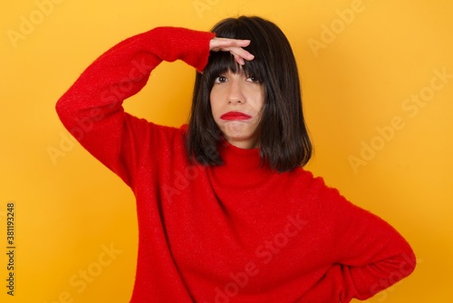 Caucasian brunette woman wearing red casual sweater isolated over yellow background, having problems, worried and stressed holds hand on forehead. © Roquillo
