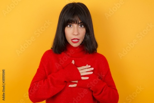 Scared Caucasian brunette woman wearing red casual sweater looks with frightened expression, keeps hands on chest, being puzzled to notice something strange, People, hush reaction and emotions.