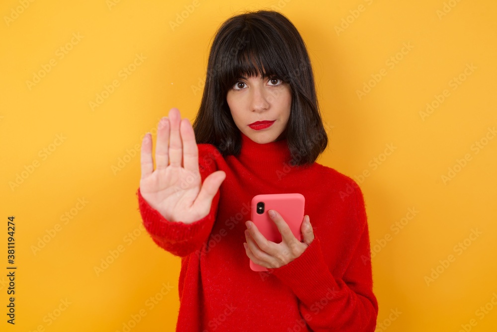 Caucasian brunette woman wearing red casual sweater isolated over yellow background using and texting with smartphone with open hand doing stop sign with serious and confident expression,