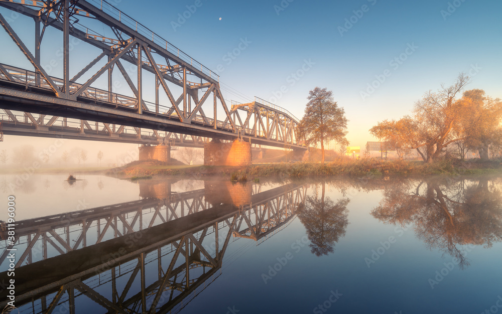 Obraz premium Beautiful railroad bridge and river in fog at sunrise in autumn. Industrial landscape with railway station, reflection in water, road, mist, trees, blue sky in foggy morning in fall. Transportation