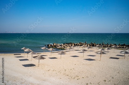 Late September empty beach in a Romanian resort at the Black Sea with straw umbrellas and rocky edges.