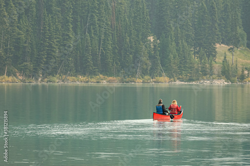 A couple canoeing along the shoreline of a calm turquoises lake forested by tall spruce trees. They wear layered clothing. They are relaxed and enjoying the smooth motion of gliding across the lake.