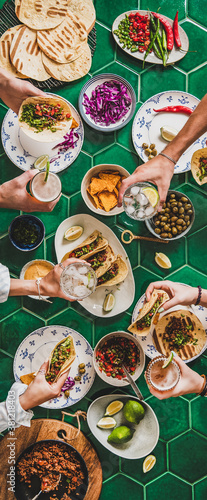 Friends home taco party. Flat-lay of Mexican traditional dishes Tacos with beef, corn tortillas , tomato salsa and peoples hands with drinks and food over green background, top view. Mexican cuisine