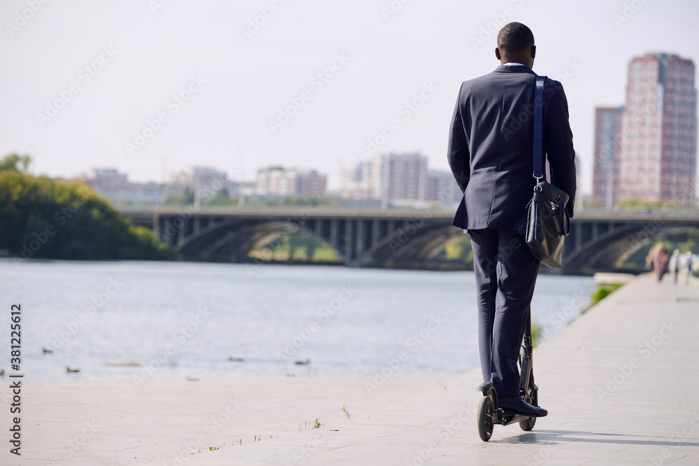 Back view of young elegant businessman in formalwear moving on electric scooter