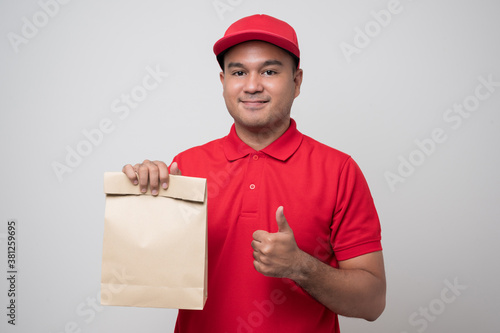 Young smiling asian delivery man in red uniform holding paper bag food delivery showing thumbs up on isolated white background.. © Chanakon