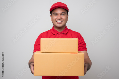 Canvastavla Young smiling asian delivery man in red uniform holding box parcel cardboard giving to customer standing on isolated white background