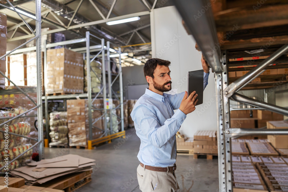 Young attractive bearded supervisor standing next to shelves in warehouse and checking on goods while holding and looking at tablet. Young attractive bearded supervisor standing next to shelves in warehouse and checking on goods while holding and looking at tablet.