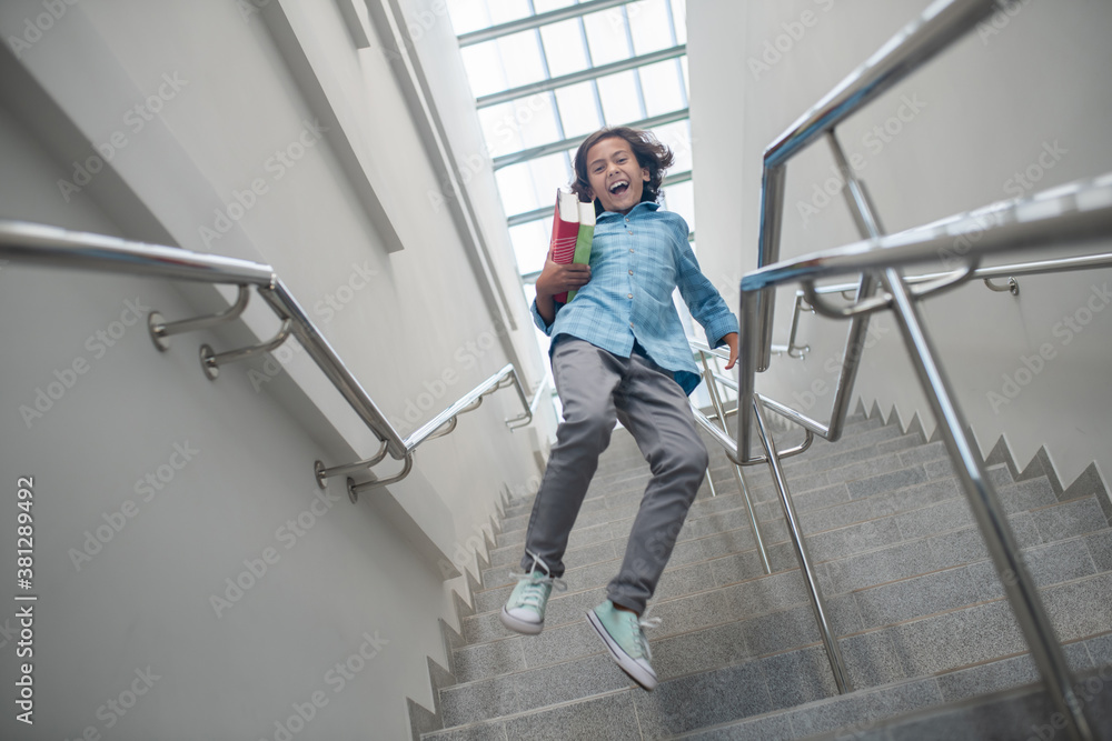 Fototapeta premium Excited schoolboy running downstairs with pile of books