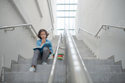 Schoolboy sitting on stairs, reading book, holding his chin