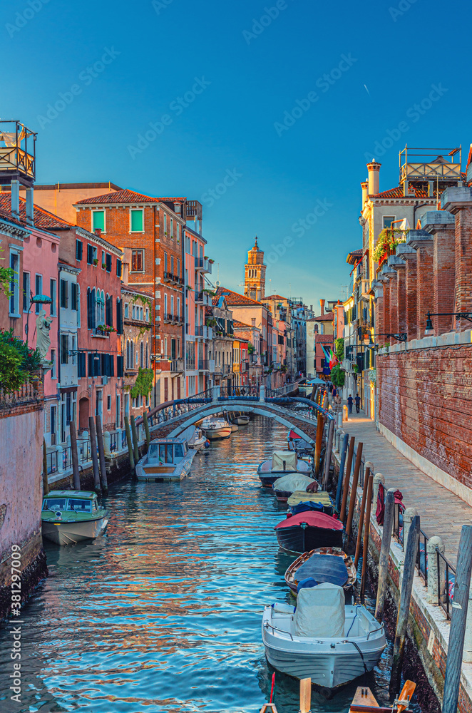 Fototapeta Venice cityscape with narrow water canal and colorful boats moored near fondamenta embankment, Veneto Region, Northern Italy. Typical Venetian view at sunset, vertical view, blue sky background
