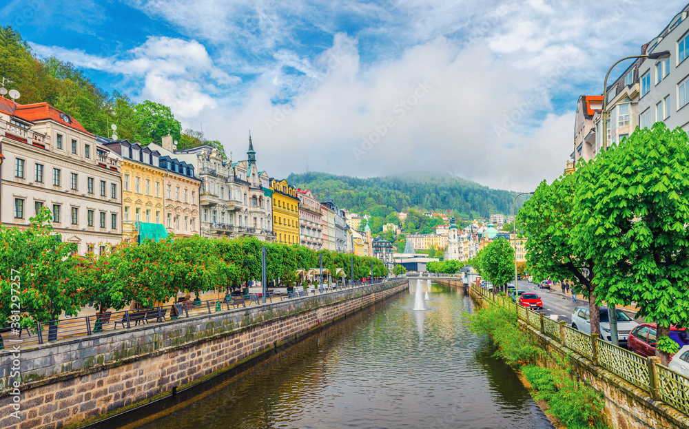 Fototapeta premium Karlovy Vary historical city centre with Tepla river pedestrian embankment, colorful beautiful buildings, Slavkov Forest hills background, West Bohemia, Czech Republic. Panorama of Karlsbad town.