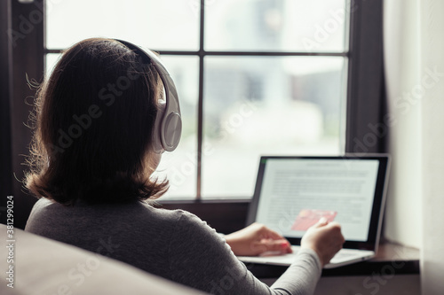 Woman holding credit card using laptop for making order sitting near window in headphones. Online shopping or booking concept.