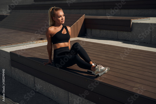 Fitness woman relaxing sitting on a bench. Cheerful sportswoman sits resting on the street at dawn in a fitness suit.