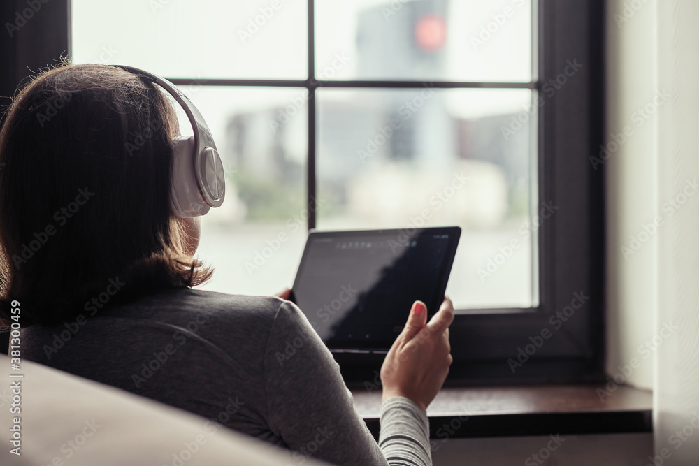 Back view of сaucasian young woman with tablet in headphones listening music sitting near window of apartments. Back view of сaucasian young woman with tablet in headphones listening music sitting near window of apartments.