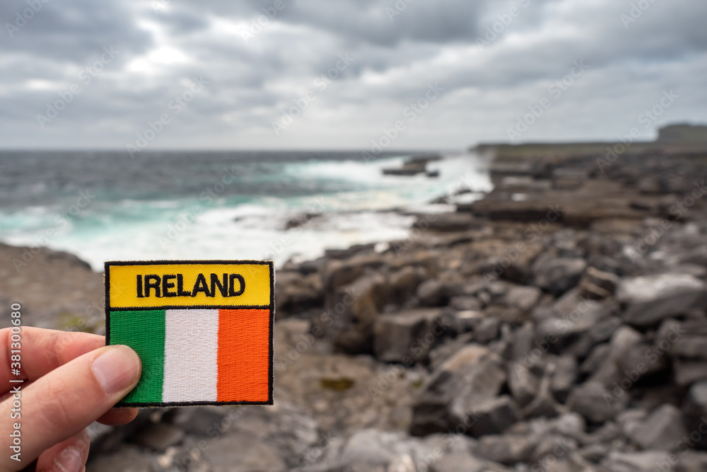 Tourist holding badge with sign Ireland and Irish National flag in focus. Rough stone coast line and Atlantic ocean out of focus. Inis mor Aran Islands, county Galway, Ireland.