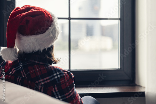 Back view of lonely woman in a red santa claus christmas hat sitting and looking at window.