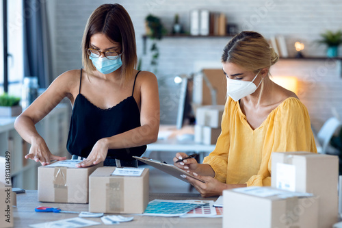 Two beautiful freelance business women seller wearing a hygienic facial mask while checking product order while packing and sealing cardboard boxes in their startup small business.
