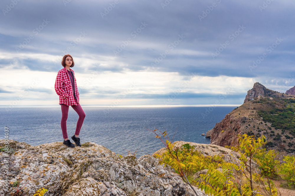 Teen tourist girl standing on mountain cliff against beautiful seascape in cloudy day. Young lady posing on background of sea, mountains and sky. Travelling and discovering distant places of Earth