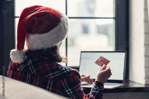 Woman in a red santa claus hat holding credit card using laptop for making order sitting near window. Christmas online shopping. Concept