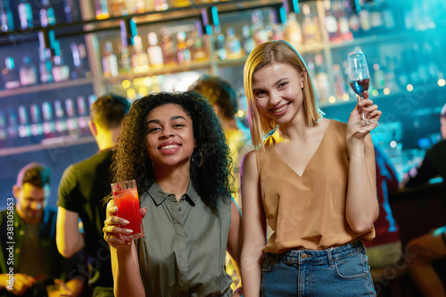 Attractive young women smiling at camera, posing with cocktail in their hands. Friends celebrating, having fun in the bar