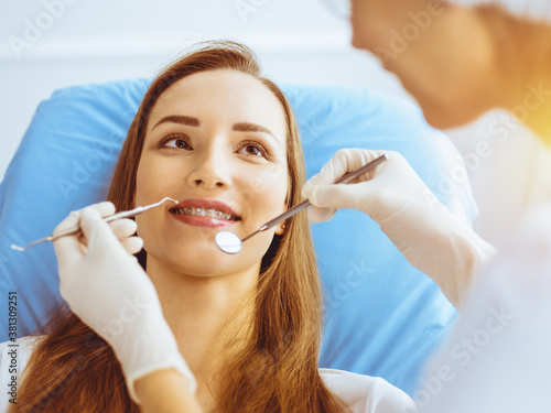 Smiling young woman with orthodontic brackets examined by dentist in sunny dental clinic. Healthy teeth and medicine concept