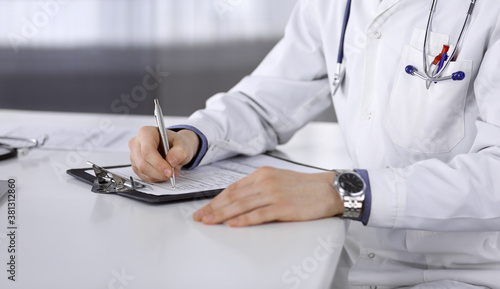 Doctor sitting and working with clipboard of medication history record in clinic at his working place, close-up. Young physician at work. Perfect medical service, medicine concept