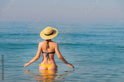 An attractive slender young Caucasian woman in a swimsuit and a straw hat stands waist-deep in sea water in the morning when there is haze over the sea