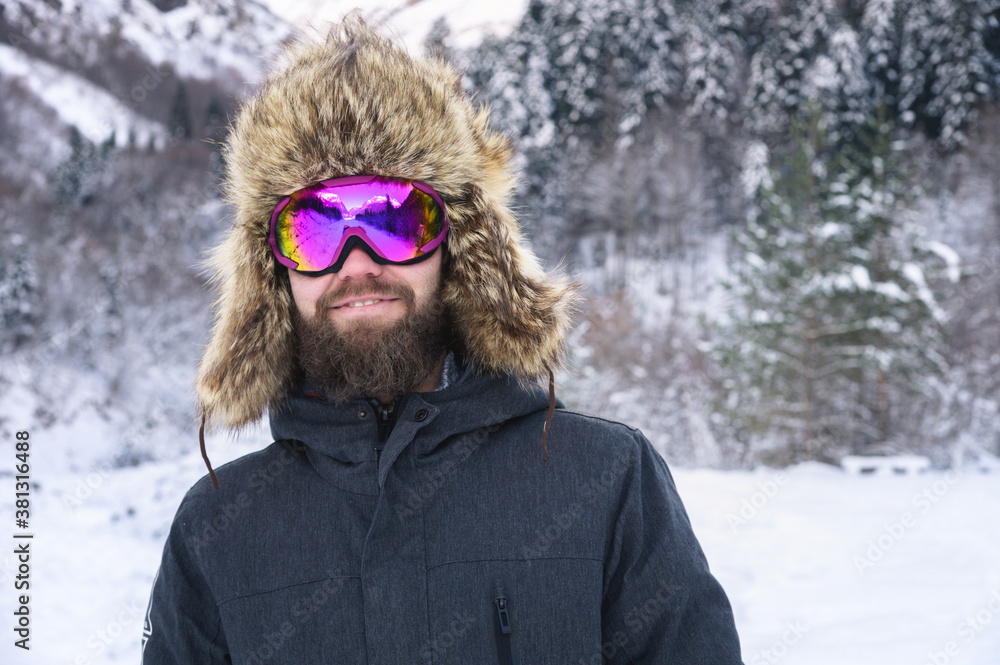 Portrait of an attractive bearded smile man in a ski mask and a fur hat against the background of a snowy forest Portrait of an attractive bearded smile man in a ski mask and a fur hat against the background of a snowy forest