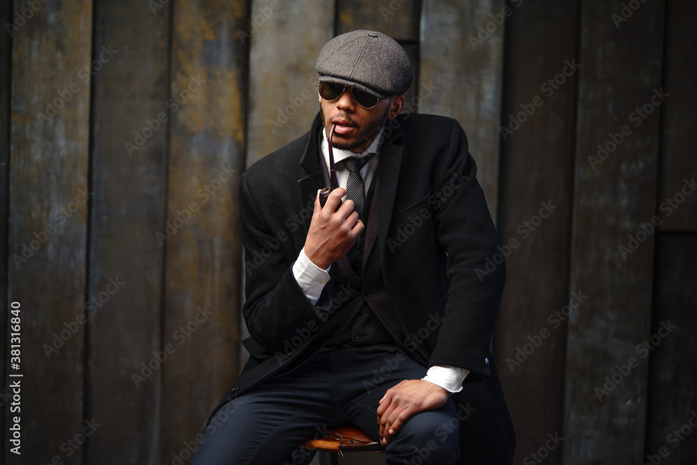 Studio shot of stylish african american man wearing coat, cap, glasses and holding smoking pipe Studio shot of stylish african american man wearing coat, cap, glasses and holding smoking pipe