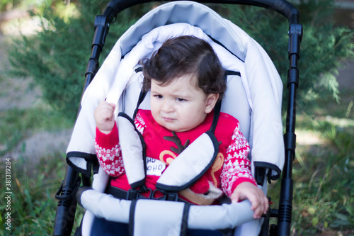 Little boy sitting in a stroller in a red sweater looks to the side and shows a cam