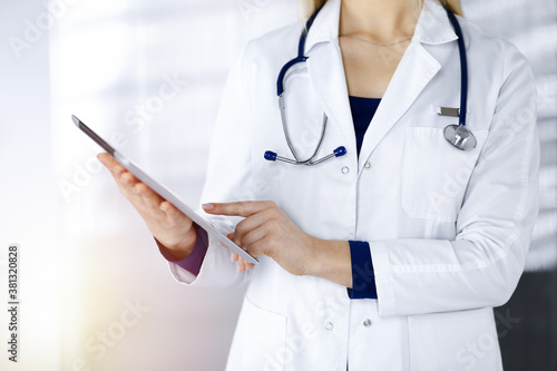 Unknown woman-doctor is holding a tablet computer in her hands, while standing in a sunny clinic cabinet. Female physician at work, close-up. Perfect medical service in a hospital. Medicine concept