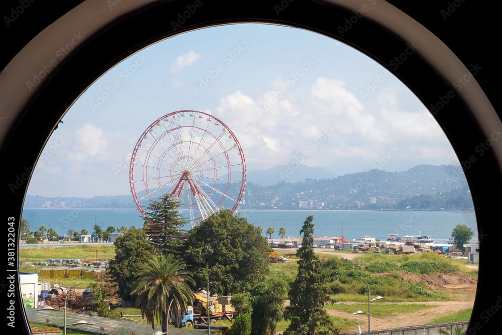 View of sea, coast and city park with ferris wheel through the round window View of sea, coast and city park with ferris wheel through the round window