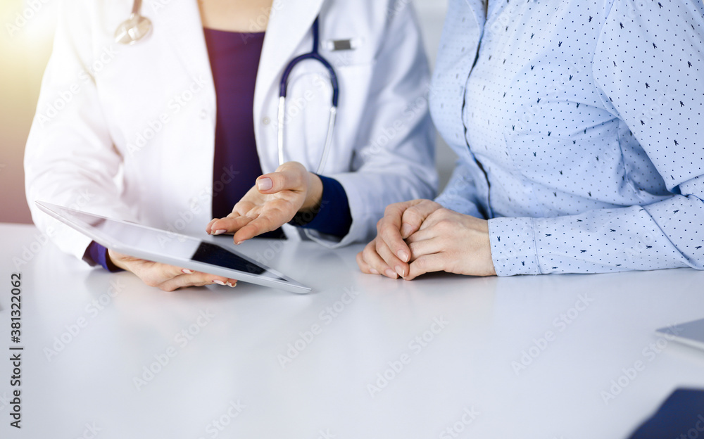 Unknown woman-doctor is showing to her patient a description of medication, while sitting together at the desk in the sunny cabinet in a clinic. Female physician is using a computer tablet and a Unknown woman-doctor is showing to her patient a description of medication, while sitting together at the desk in the sunny cabinet in a clinic. Female physician is using a computer tablet and a