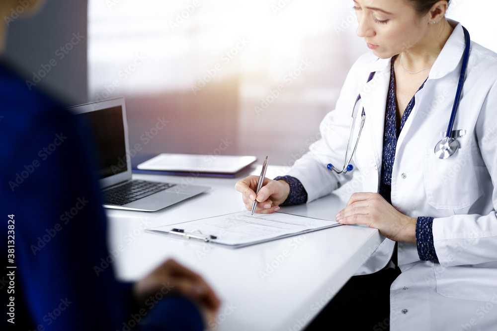 Unknown woman-doctor is writing some medical recommendations to her patient, while they are sitting together at the desk in the sunny cabinet in a clinic. Physician is using a clipboard, close-up Unknown woman-doctor is writing some medical recommendations to her patient, while they are sitting together at the desk in the sunny cabinet in a clinic. Physician is using a clipboard, close-up