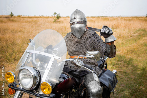 A medieval knight in chainmail and a helmet with a sword in his hands sits on a motorcycle.