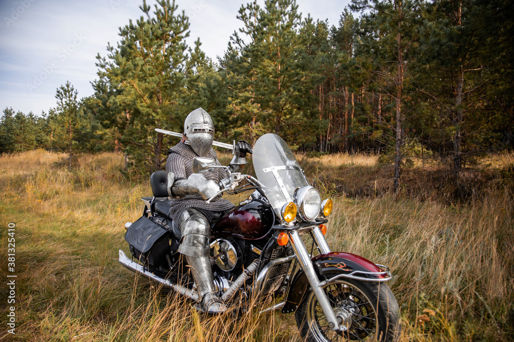 A medieval knight in chainmail and a helmet with a sword in his hands sits on a motorcycle. A medieval knight in chainmail and a helmet with a sword in his hands sits on a motorcycle.