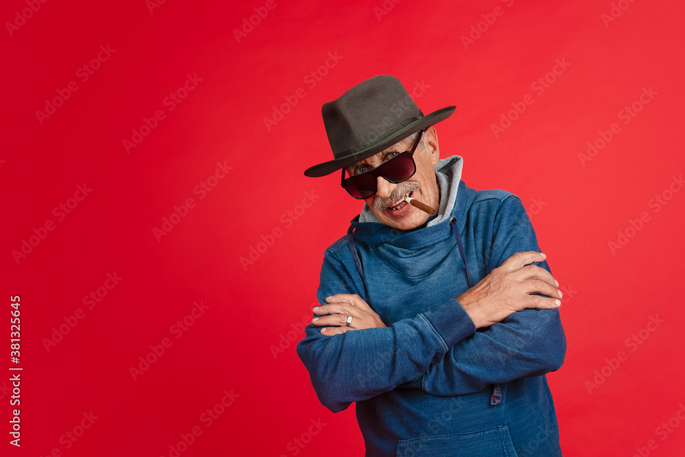 Smiling, smoking. Portrait of senior man in stylish eyewear and hat isolated on red studio background. Tech and joyful elderly lifestyle concept. Trendy colors, forever youth. Copyspace for your ad. Smiling, smoking. Portrait of senior man in stylish eyewear and hat isolated on red studio background. Tech and joyful elderly lifestyle concept. Trendy colors, forever youth. Copyspace for your ad.
