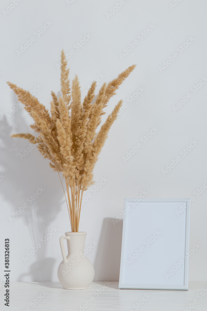 Plakat A vase with yellow spikelets and a white photo frame on the table. Light stylish background. Preparation for inscriptions, copy space.