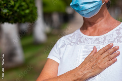 A senior woman white dressed  wearing surgical mask due to coronavirus standing and greeting with new recommended gesture by World Health Organization, with the hand on the heart