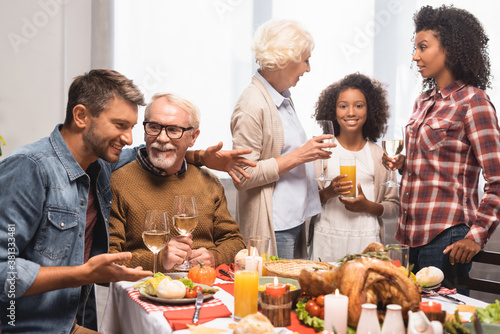 excited multicultural family talking while celebrating thanksgiving at table with festive dinner