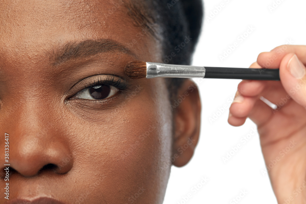 beauty, cosmetics and people concept - close up of face of beautiful young african american woman and hand with make up brush applying eye shadow over white background