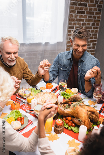 joyful multicultural family holding hands during dinner on thanksgiving holiday