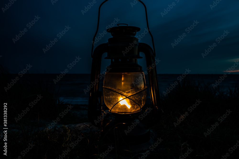 Old classic oil lantern burning with an orange flame by the ocean at dusk.