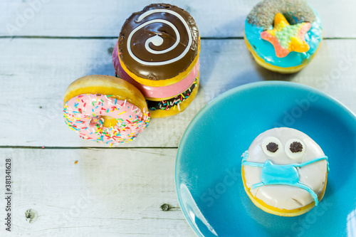 top view of a handmade donuts with face mask due to covid pandemic on a blue plate and colorful donuts on wooden table